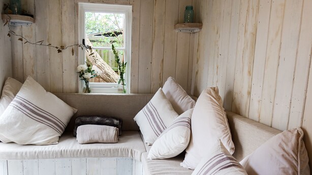 A cozy room with a wooden bench and a cushioned bench, both with pillows. A window with a view of greenery is visible. -  The Cook's Cottage: a coastal oasis w/outdoor kitchen, garden & cinema nr beach (Whitstable)