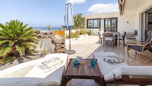 A sunlit patio with lounge chairs, a table with drinks, and a view of the sea. - Stunning beach view, heated pool, perfect house for comfort and relaxation   (Urbanización Famara)