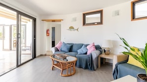 A spacious living room with a sofa, a coffee table, and a side table. There is a large window and a door leading to an outdoor area. - Stunning beach view, heated pool, perfect house for comfort and relaxation   (Urbanización Famara)