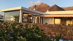 A modern house with a stone wall, large windows, and a mountain in the background. - Stunning beach view, heated pool, perfect house for comfort and relaxation   (Urbanización Famara)