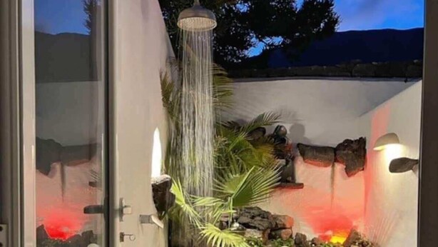 A modern outdoor shower area with a waterfall feature, surrounded by rocks and palm plants. - Stunning beach view, heated pool, perfect house for comfort and relaxation   (Urbanización Famara)