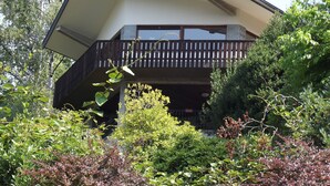 Een modern huis met een schuin dak, een balkon met houten leuningen en weelderige begroeiing eromheen.