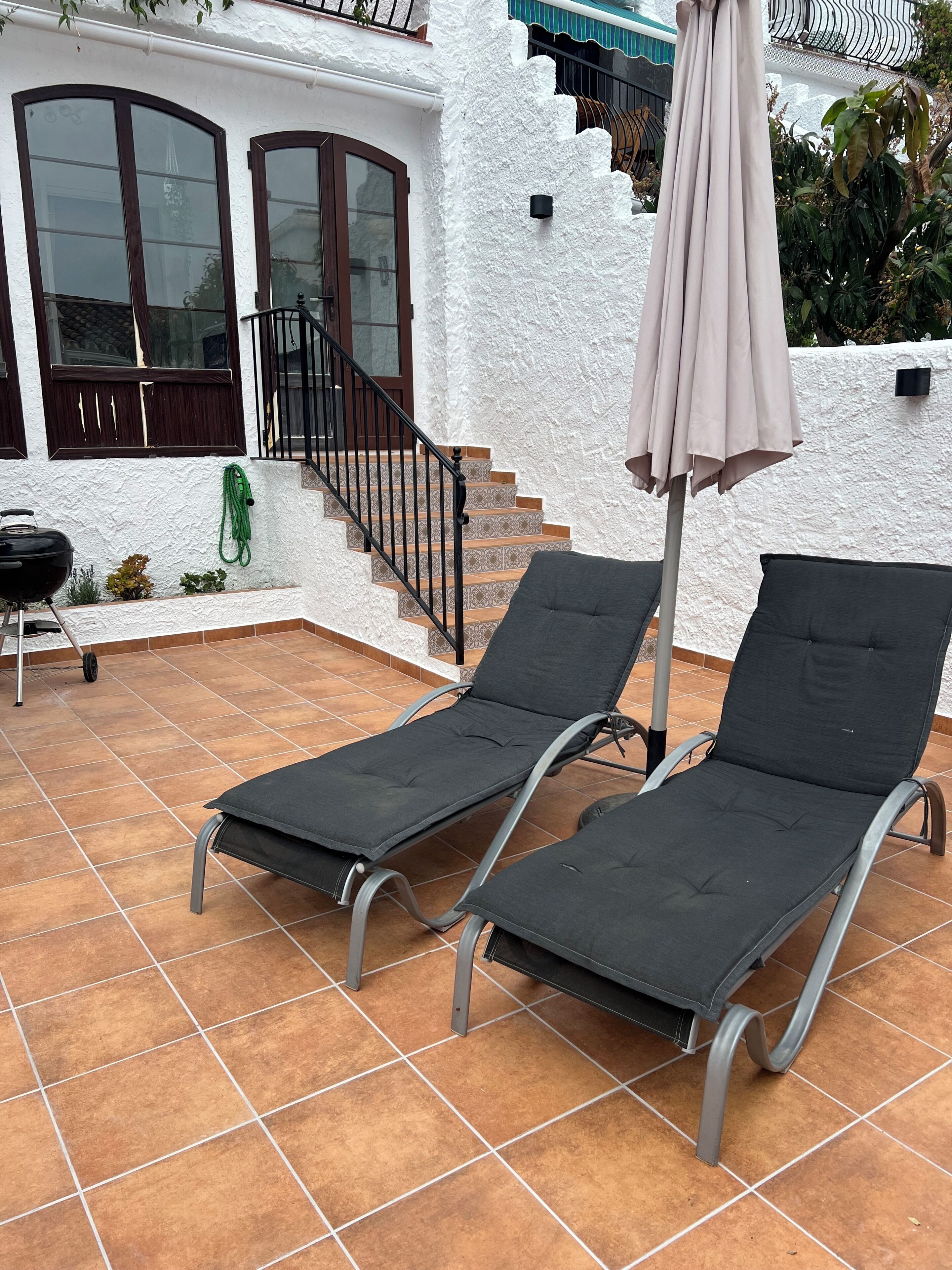A patio with two lounge chairs, a barbecue grill, and a staircase leading to a balcony.