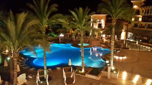 Une vue nocturne d’une piscine entourée de palmiers et de chaises longues, avec un bâtiment en arrière-plan.