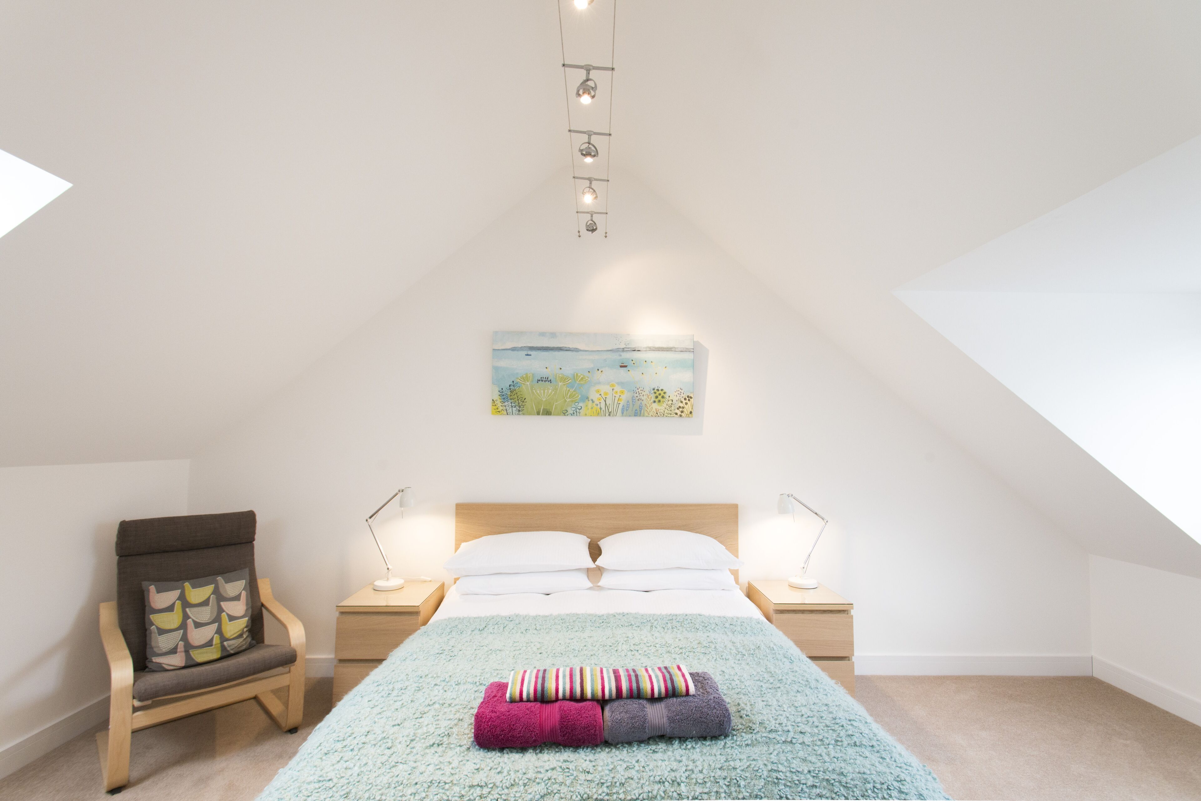 A bedroom with a sloped ceiling, a bed with white linens, a chair with a patterned cushion, and a painting on the wall.