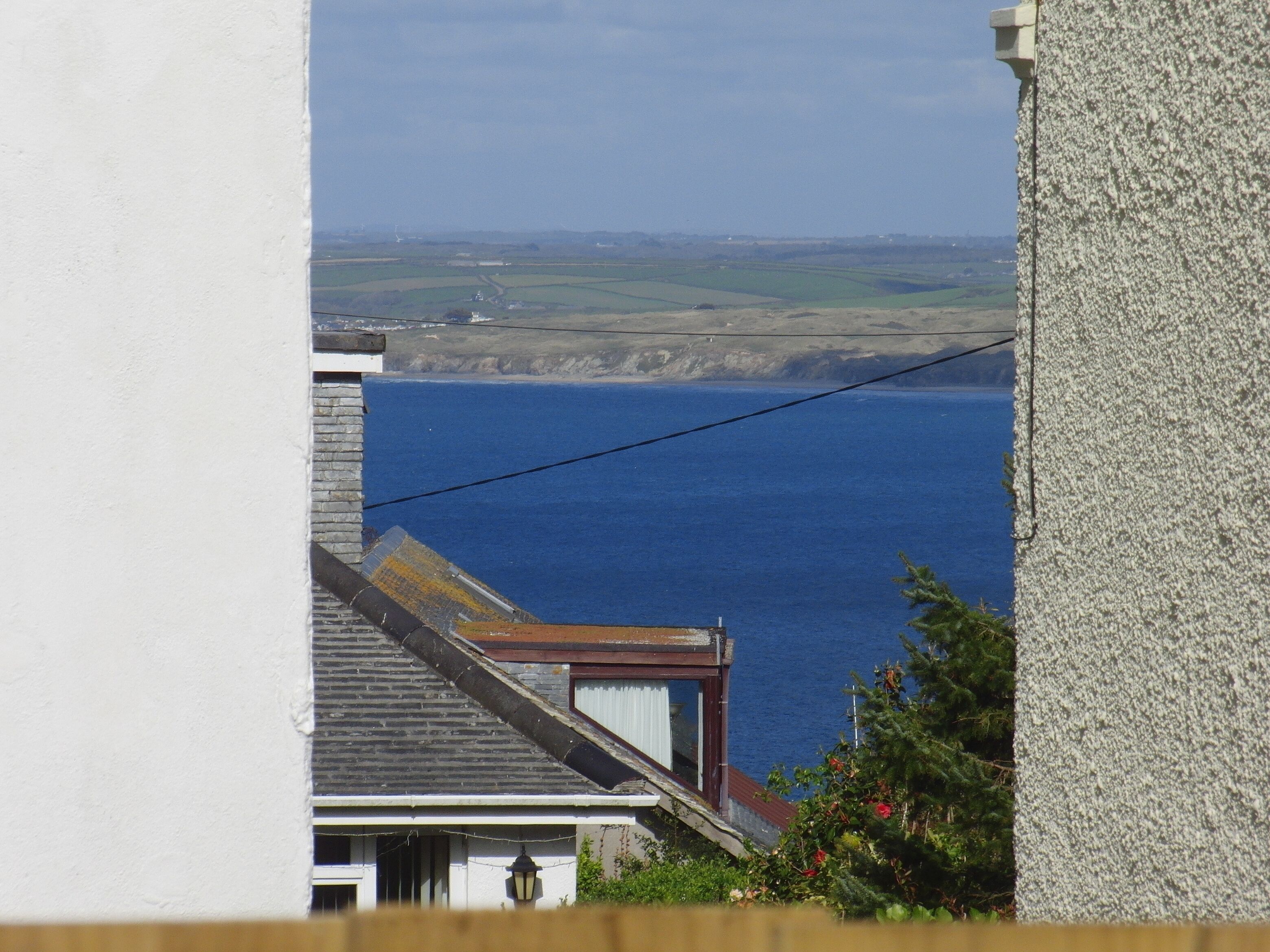 A coastal view from a building with a visible power line, a house with a sloped roof, and a clear view of the sea and distant landscape.