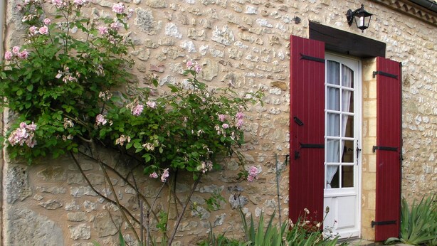 A stone building with a red door and shuttered window, surrounded by greenery and flowering plants. - In the center of Périgord  Noir (SARLAT 20 Km)- LE COUX et BIGAROQUE.  Périgourdine house with  swimming pool, beautiful panorama on valley, on property of 6 ha. (Coux Et Bigaroque-Mouzens)
