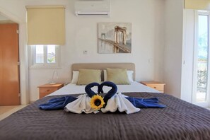 A hotel room with a bed, a nightstand, a window with a view, an air conditioner, and a painting on the wall.