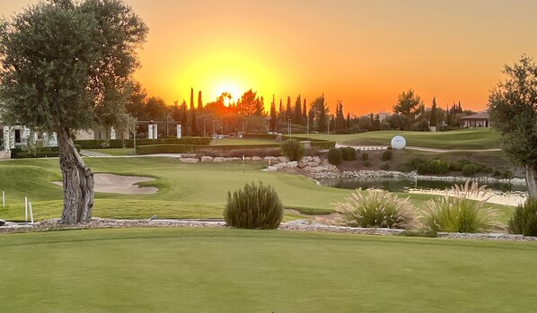 A golf course with a sunset view, well-maintained grass, sand bunkers, and a pond.
