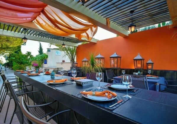 An outdoor dining area with tables set for two, surrounded by chairs, and a pergola overhead.