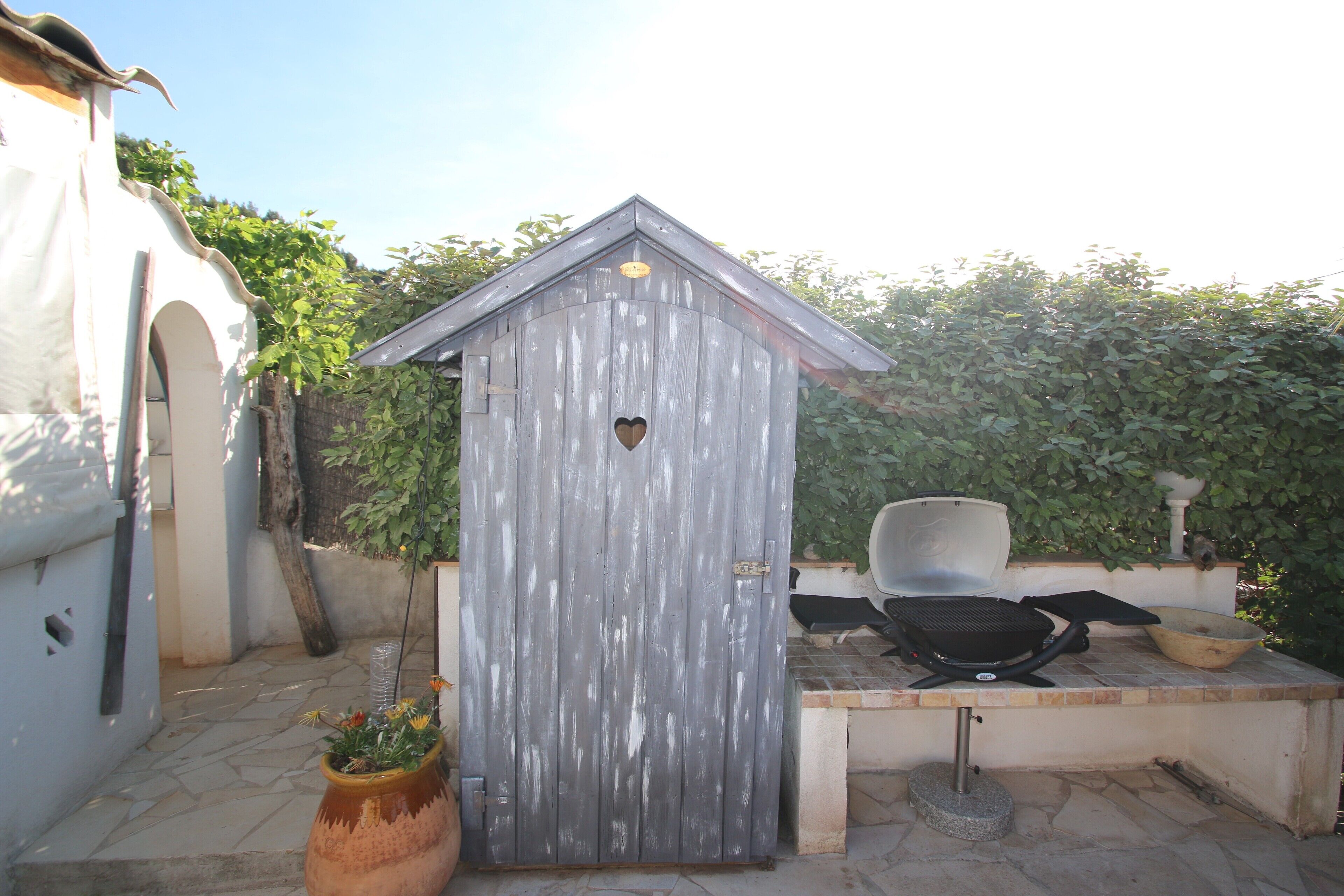 Un petit abri en bois avec une fenêtre en forme de cœur, un barbecue et une plante en pot.