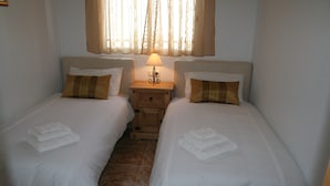 Ein Zweibettzimmer mit zwei Betten, jeweils mit weißer Bettwäsche und braunen Kissen, einem Nachttisch mit Lampe und einem Fenster mit durchsichtigen Vorhängen.