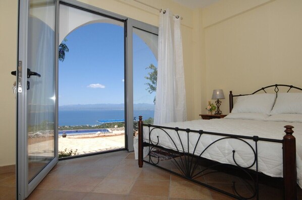 A bedroom with a bed, a nightstand, a lamp, and a view of the sea through a sliding glass door.