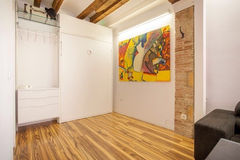 Ein Zimmer mit Holzboden, einem weißen Kleiderschrank, einem Glastischchen mit Hüten und einem farbenfrohen abstrakten Gemälde an der Wand.