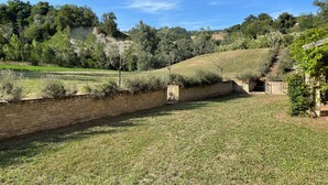 Une cour verdoyante, avec un mur de briques, un escalier en pierre et une vue sur une colline boisée.