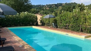 Une piscine rectangulaire à l’eau d’un bleu limpide, entourée d’une terrasse en briques. On y trouve deux chaises longues noires et une table avec un parasol noir. La piscine est bordée d’un petit muret en briques, et un luxuriant décor de verdure et d’arbres se trouve en arrière-plan.
