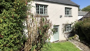 Une maison à deux étages, à l’extérieur blanc et aux encadrements de fenêtres verts, entourée de verdure et bordée d’un chemin en pierre.
