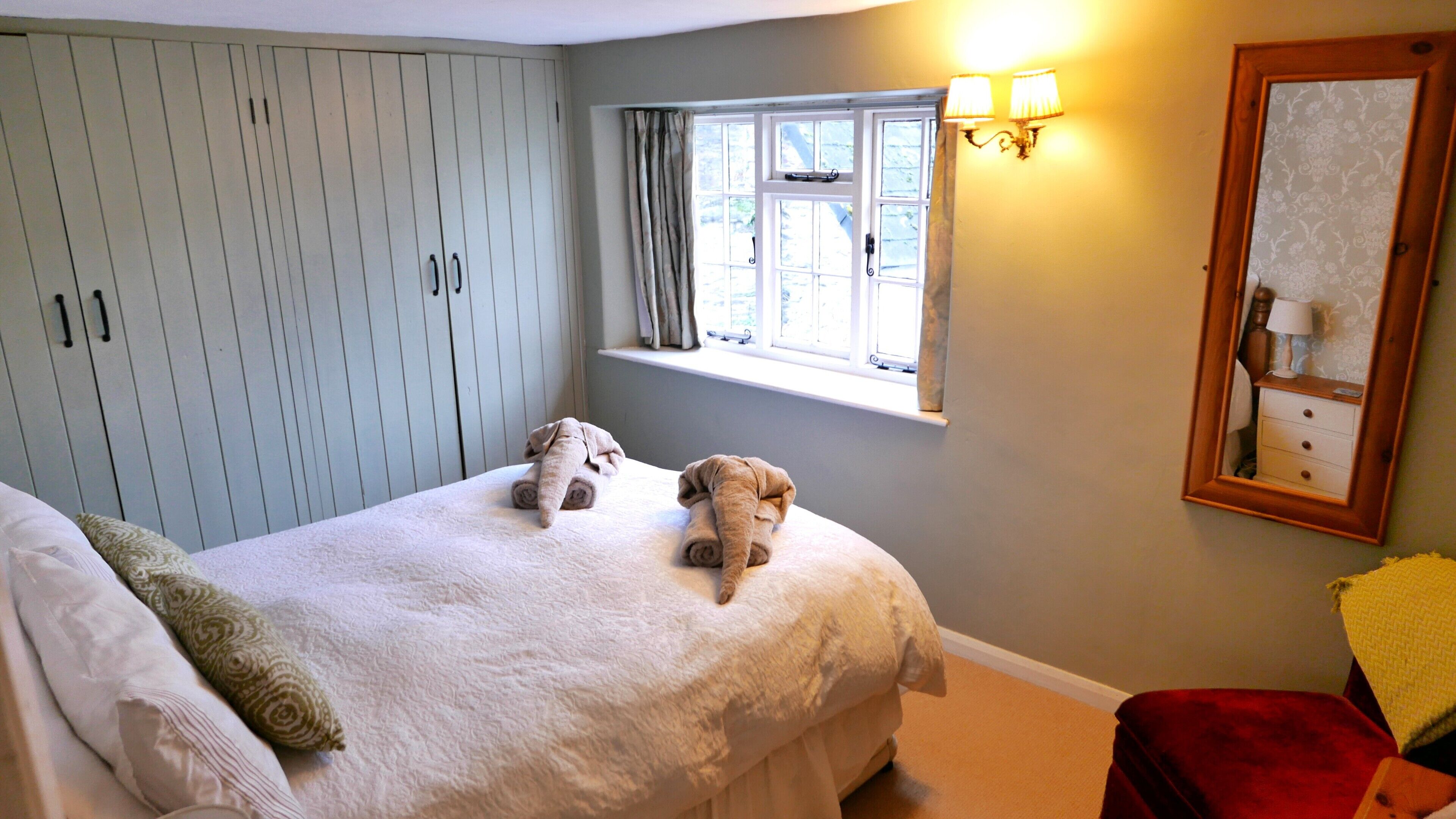 A bedroom with a bed, two rolled-up towels, a window with curtains, a nightstand with a lamp, and a mirror on the wall.