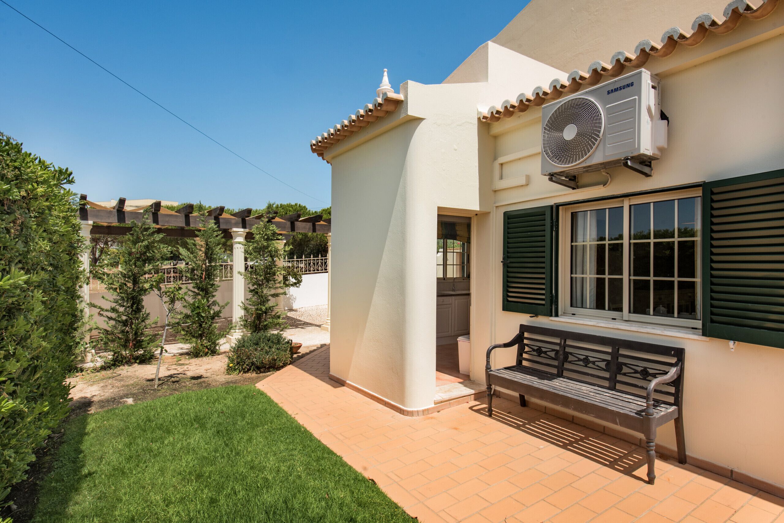 Edifici amb façana blanca, un aparell de aire condicionat Samsung fixat a la paret, una banca a l'exterior i un jardí ben cuidat amb gespa i arbustos.