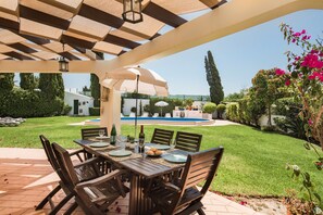 An outdoor patio with a table set for dining, surrounded by chairs, and a pool in the background.