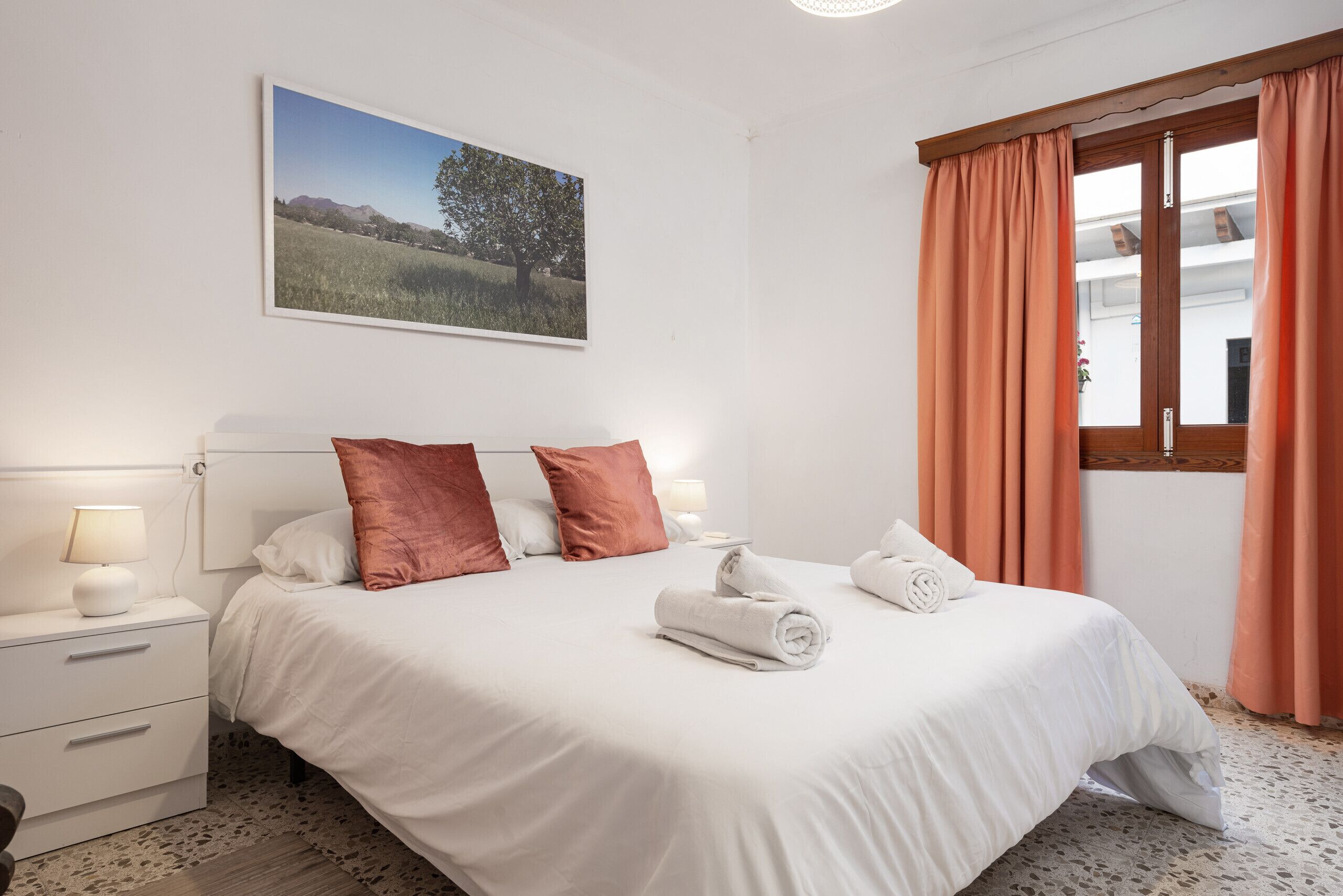 Un lit bien fait, avec du linge de lit blanc et deux oreillers, une table de chevet avec une lampe, et une fenêtre ornée de rideaux orange.