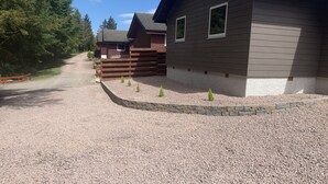 Une allée de gravier mène à un chalet en bois bordé de pierres, et à une maison moderne à la façade en pierre.
