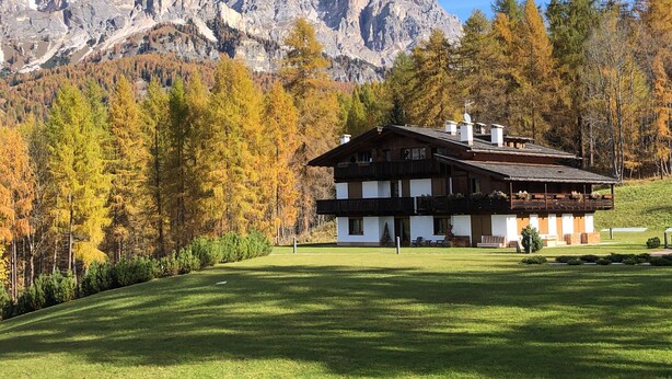 Un bâtiment de style chalet, doté d’un toit en pente et d’une façade en bois, entouré d’une pelouse bien entretenue et d’arbres aux couleurs automnales, avec une chaîne de montagnes en arrière-plan.