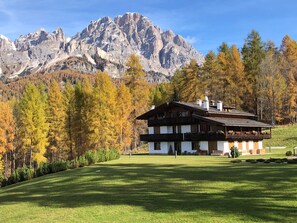 A chalet-style building with a sloped roof and wooden exterior, surrounded by a well-maintained lawn and autumn-colored trees, with a mountain range in the background.