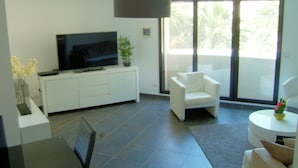 A modern living room with a flat-screen TV, a white sofa, a small round table, and a large window with a view of greenery. - Luxury apartment, perfect for beach and exploring Juan-les-Pins and Antibes,  (Antibes)