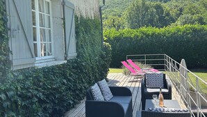 Una terraza de madera con mobiliario exterior, una casa con contraventanas blancas y vistas a colinas verdes.