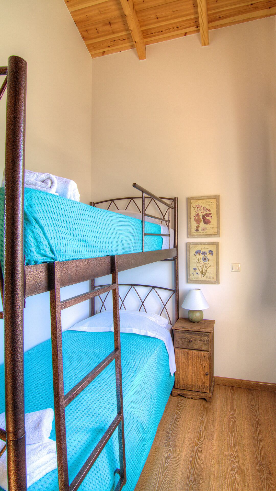 Una cama litera con estructura de madera, una mesita de noche con lámpara, y dos ilustraciones botánicas enmarcadas en la pared.