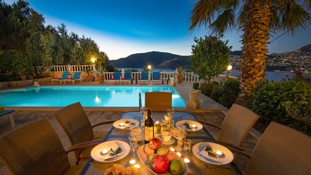 Mesa para dos preparada con platos de fruta, una botella de vino y velas, con vista a una piscina y un paisaje urbano al anochecer.