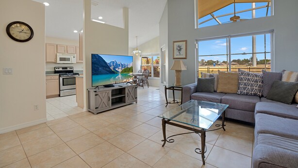 A spacious living room with a large flat-screen TV, a glass coffee table, and a comfortable sofa. A kitchen with modern appliances is visible in the background.