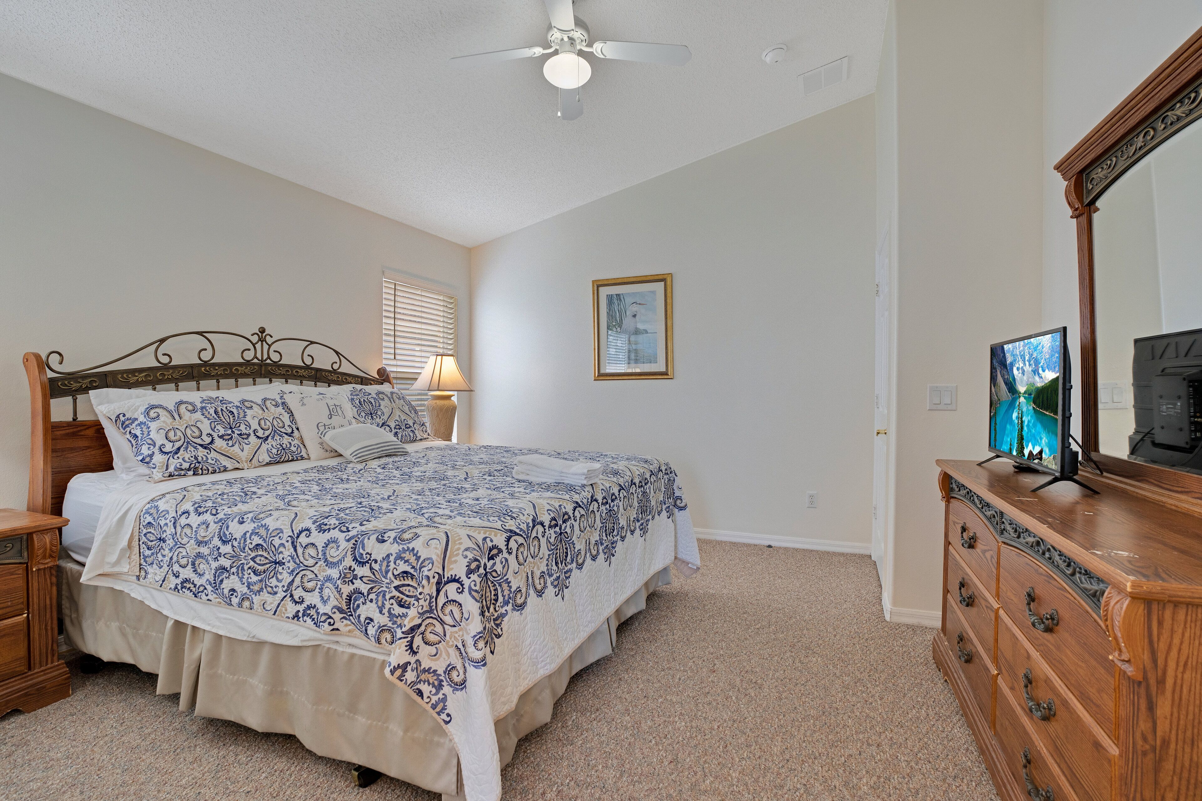 A bedroom with a bed, wooden dresser, ceiling fan, and a framed picture on the wall.