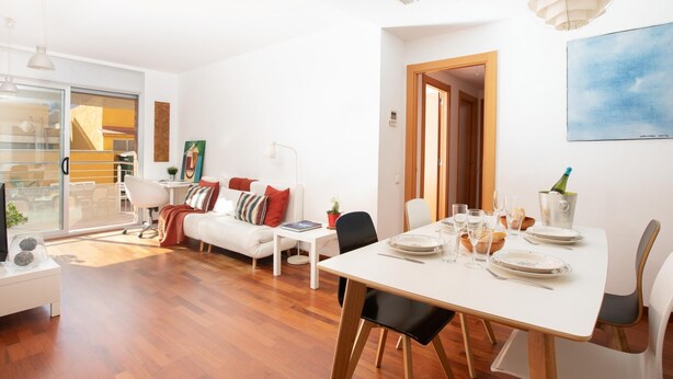 Una sala de estar moderna con comedor, que presenta una mesa blanca dispuesta para cuatro personas, un sofá con cojines de colores y un ventanal con vistas al exterior.