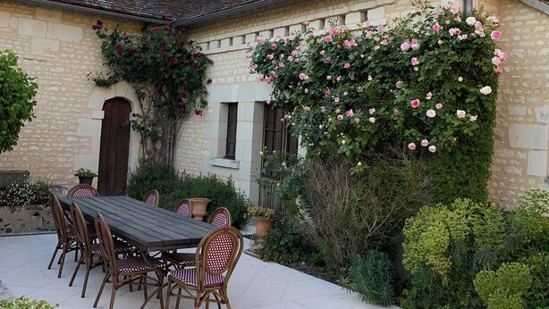 Un edificio de piedra con tejado de tejas, un comedor con mesa y sillas, y un jardín con plantas en flor.