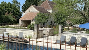 Una casa de piedra con techo de tejas, piscina y un patio vallado con sillas de descanso.