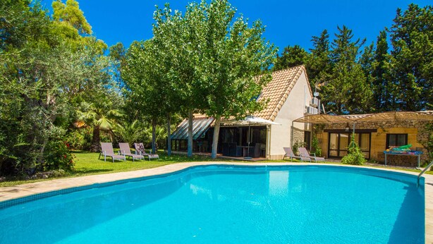 A swimming pool with lounge chairs, a covered patio, and a building with a tiled roof.