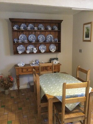 Un coin repas avec une table et des chaises en bois, un buffet en bois orné d’assiettes bleues et blanches, et un petit vase avec des fleurs rouges.