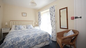 A bedroom with a bed, a nightstand, a wicker chair, a mirror, and a window with curtains.