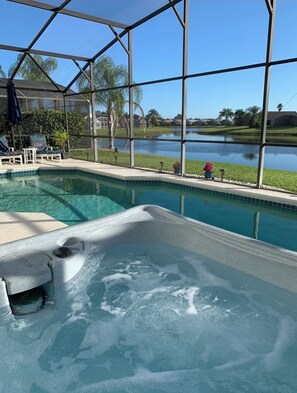Piscina com hidromassagem, rodeada por uma área coberta com cadeiras e vista para um lago e casas.