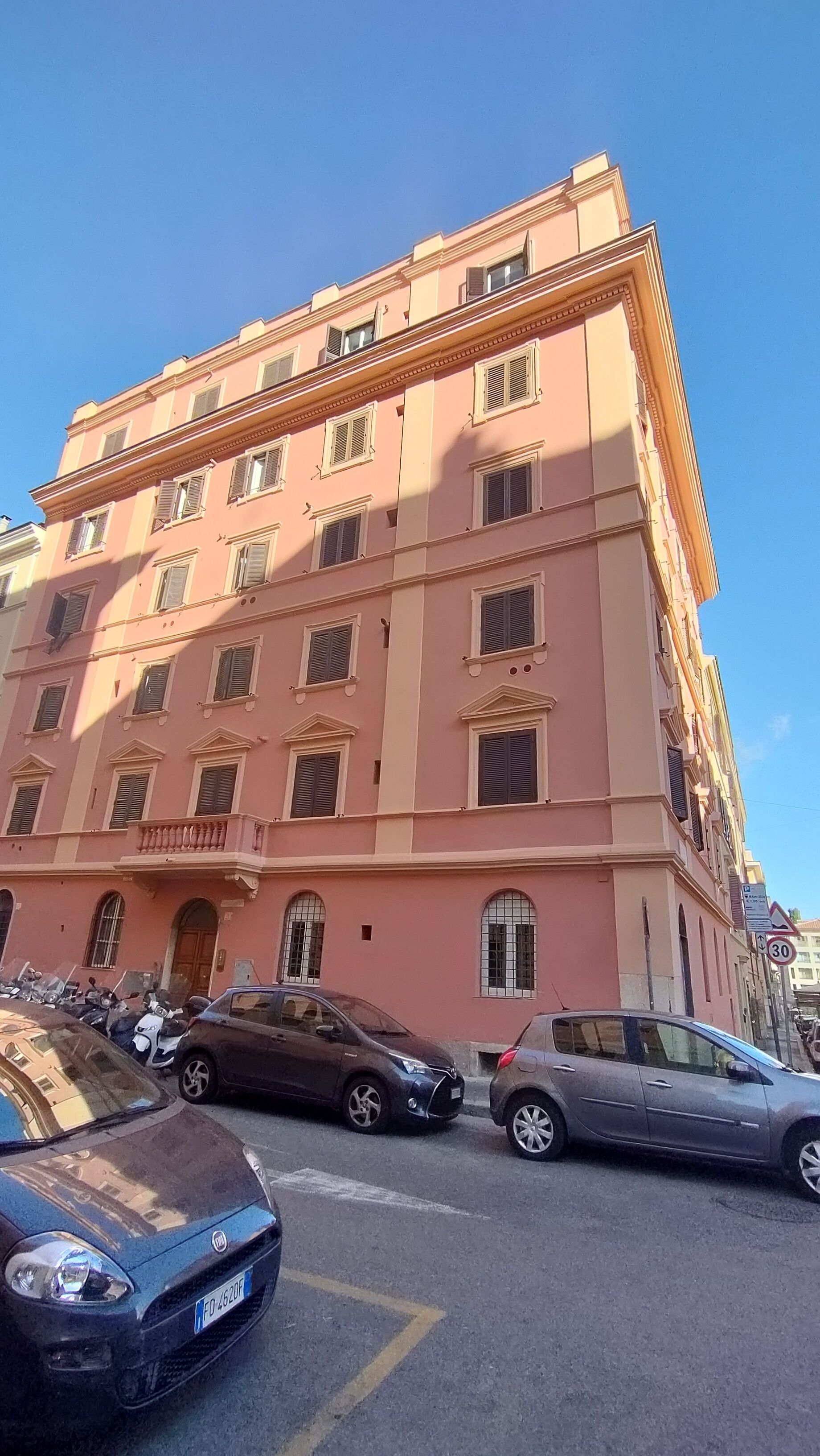 A multi-story building with a pink facade, several windows with shutters, and a parking area with cars.