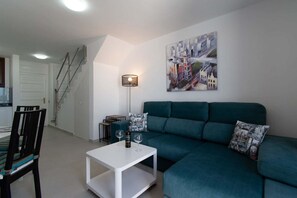 Een moderne woonkamer met een blauwgroene bank, een witte salontafel en een schilderij van een stadsgezicht.