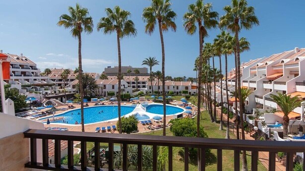 A resort with a central swimming pool surrounded by palm trees, multiple balconies, and a clear blue sky.