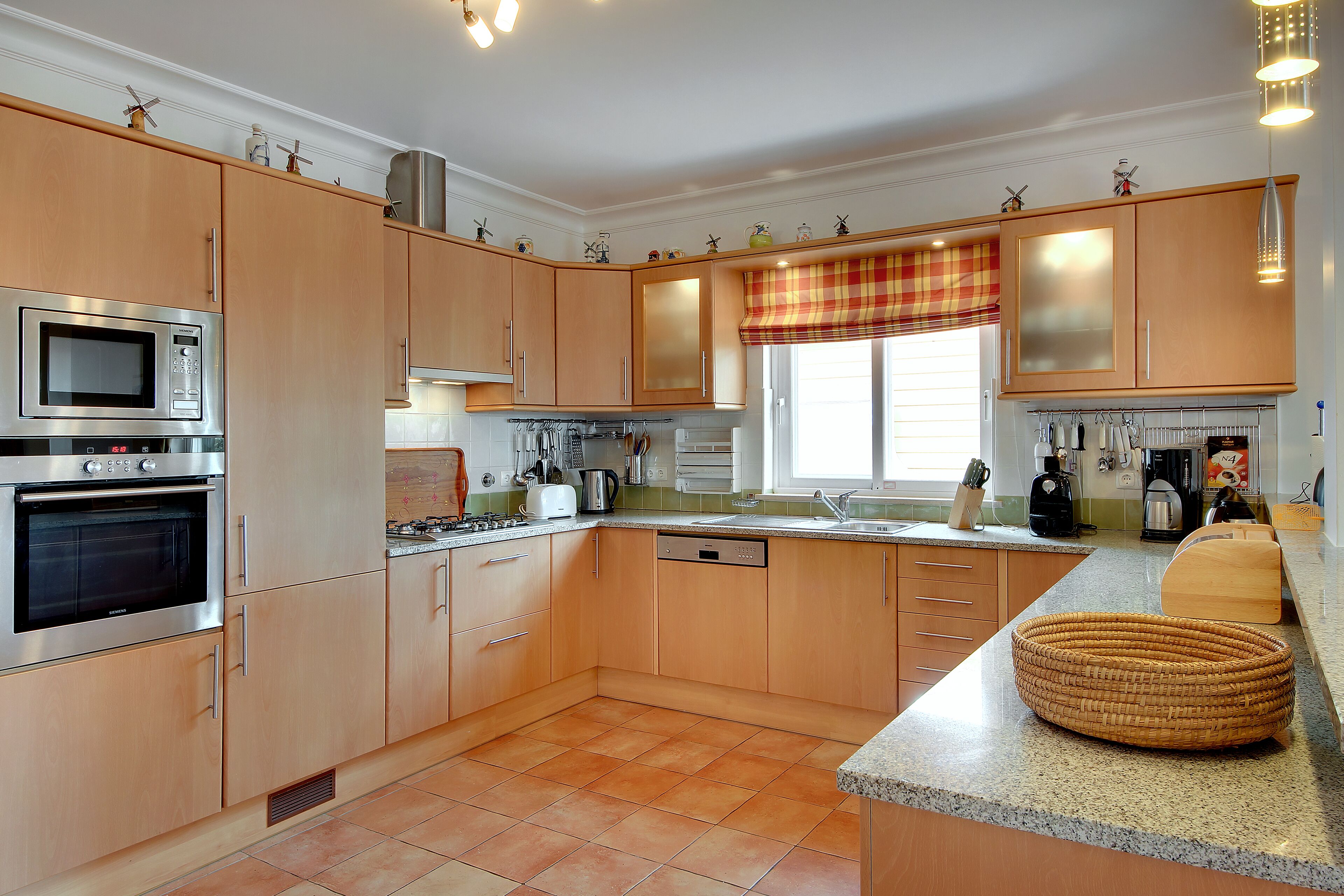 A modern kitchen with wooden cabinets, stainless steel appliances, and a large window with a red and yellow checkered valance.