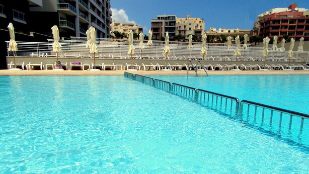 Ein Swimmingpool mit weißen Liegestühlen und Sonnenschirmen, umgeben von Gebäuden.