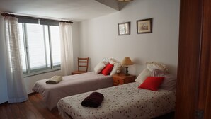 Una camera da letto con due letti singoli, una sedia in legno, un comodino con una lampada e una finestra con tende.