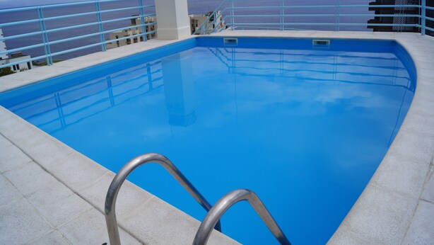 Una piscina con una scaletta metallica, vista sul mare e un edificio sullo sfondo.
