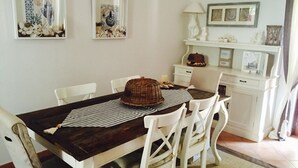 Un comedor con una mesa de madera, sillas blancas y un mantel a rayas. Se observan obras de arte enmarcadas en las paredes y un armario blanco con objetos decorativos.