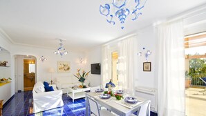 A bright living room with a white sofa, a dining table set with blue and white plates, and a blue-tiled floor. - Il canto delle sirene,  in the heart of Amalfi Coast, parking Property overview (Conca dei Marini)